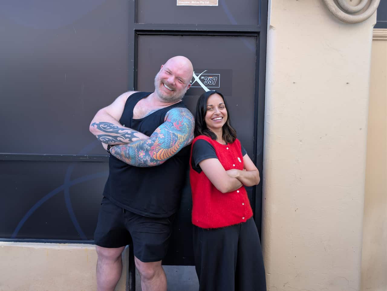 Glenn McNamara, a middle-aged white man with tattoos, and Farz Edraki, an Iranian Australian woman in her 30s wearing a red shirt, stand back to back in front of a dark building.