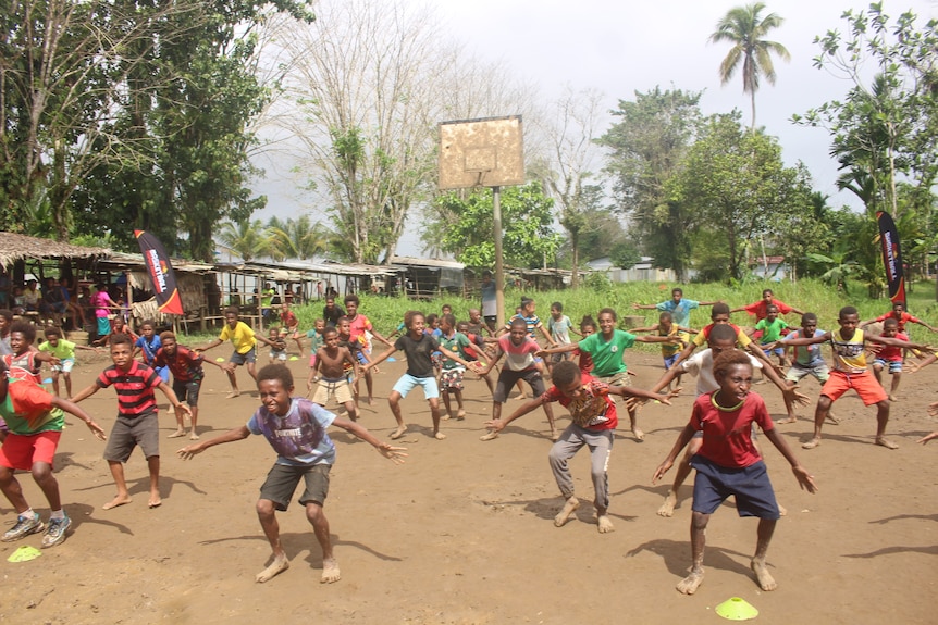 A group of young people stand on a dirt court with a basketball hoop in the background doing training exercises 