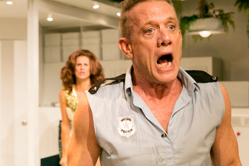 A close-up of Simon Burke, in his mid-50s, sleeveless, yelling on stage. A man in a wig is upset, blurred, behind him.