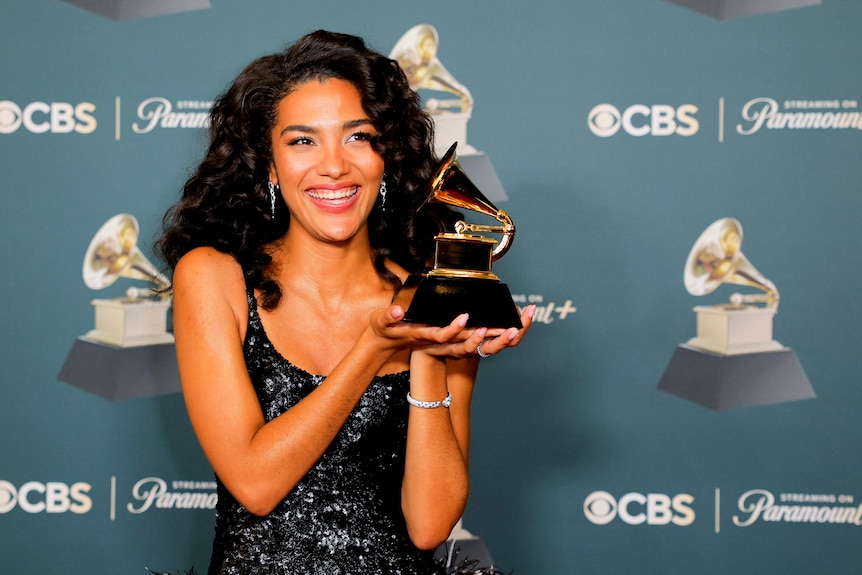 Olivia Dean holds up a grammy award against a media wall