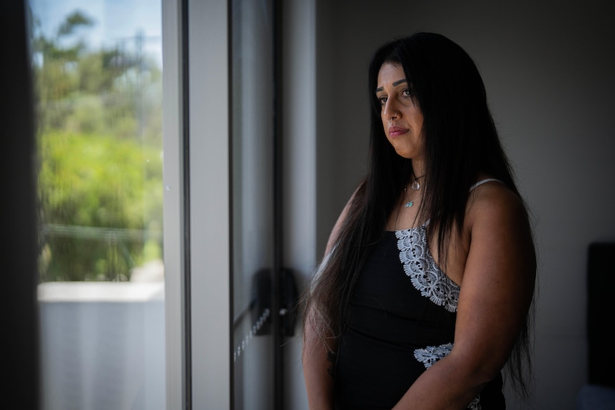 Krystle has a worried expression as she looks out of a window from a dimly lit room.