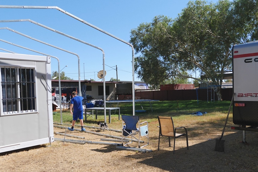 A kid walking through a backyard with a caravan and donga and poles.