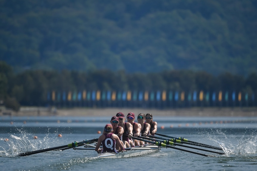 A wide shot of a women's eight rowing crew during a race. They are wearing maroon and the cox has a 'Q' on her back