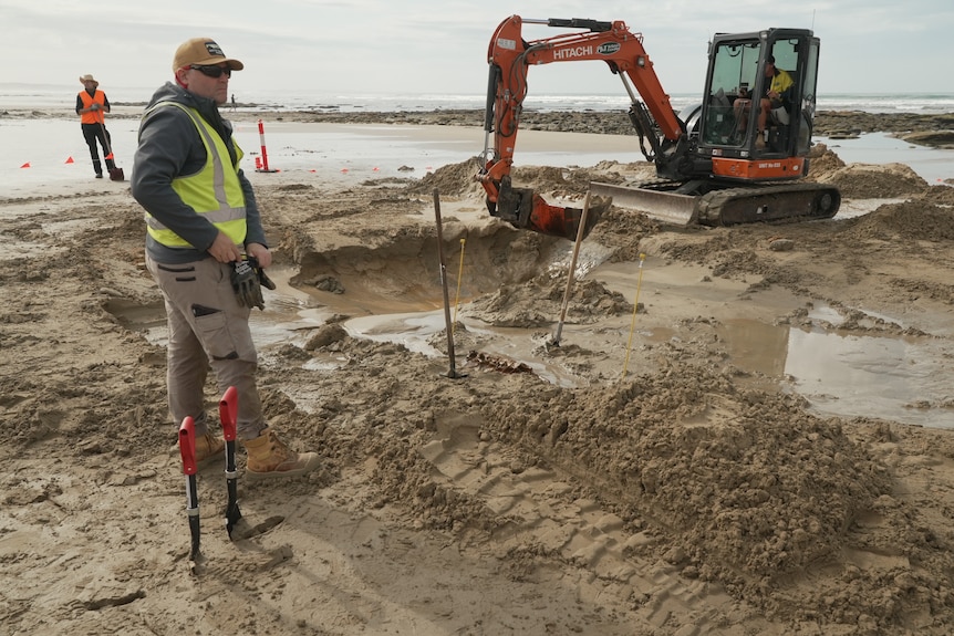 Man in high vis standing on beach in front of a digger cooping up sand.