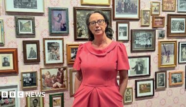 A woman in a pink dress stands against a backdrop of small photographs on a pink and white wall