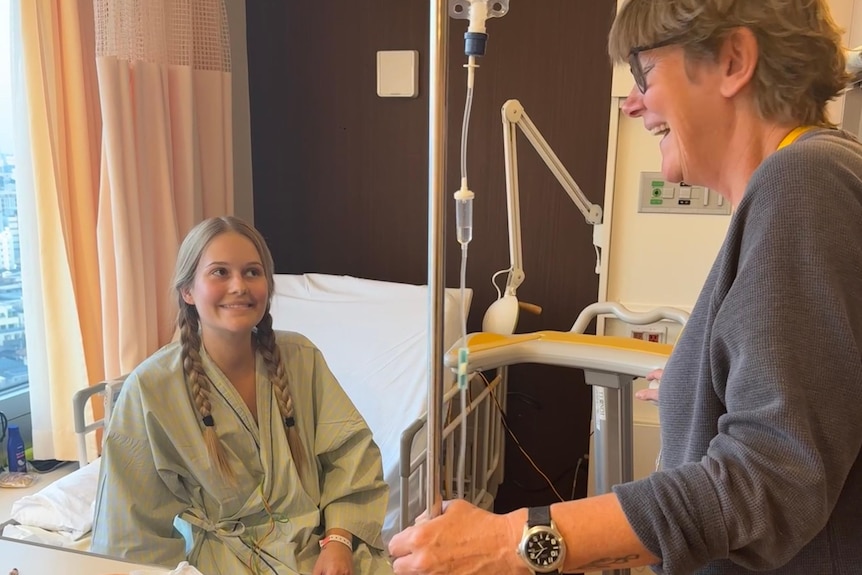 A young woman with piggy-tail blonde plaits looks up from a hospital bed at her laughing mother.