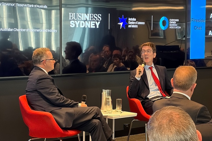 A man talks while sitting on a stage with another man.
