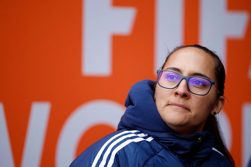 Woman with dark hair tied back off her face and glasses looks into distance