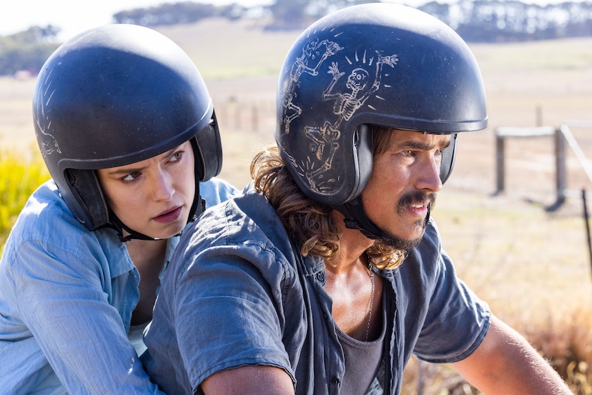 A man and a woman wearing open-face helmets, riding a motorbike through the countryside.
