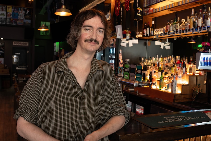 A man with brown hair and a moustache wearing a brown shirt in a dark room with alcohol bottles behind