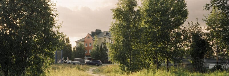Apartment building partially hidden by tall green trees, with a few cars parked nearby and a grassy path in the foreground under a cloudy sky.