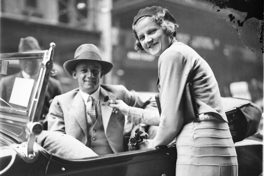 A young Don Bradman dressed in a suit and hat, sitting in a car. Young woman pins a poppy flower to his jacket.