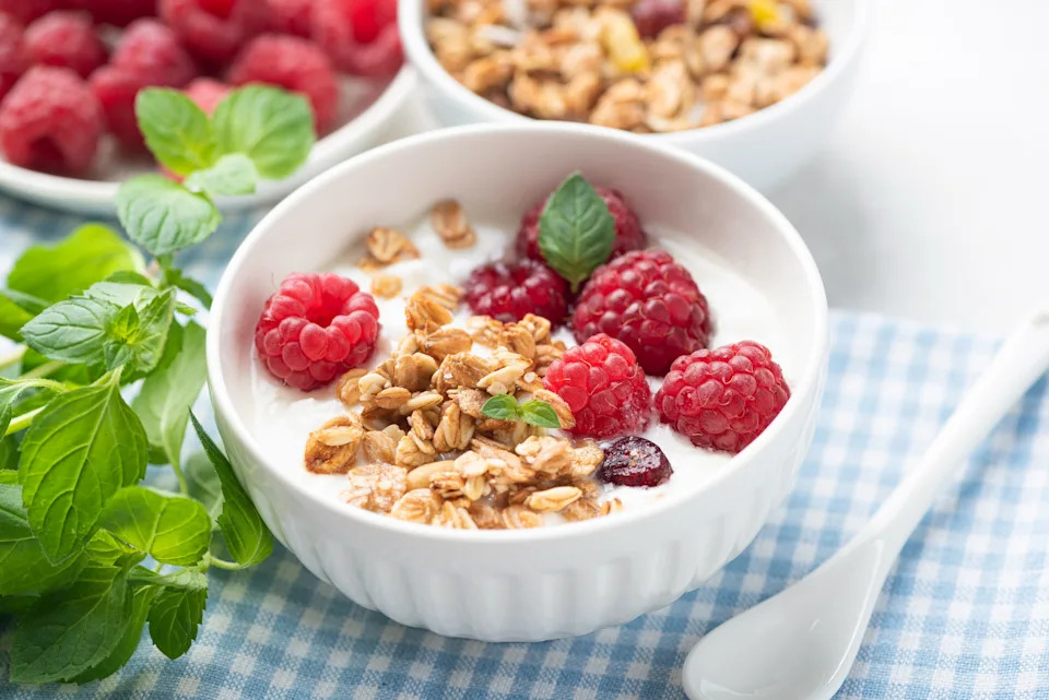 A bowl of yogurt topped with granola and raspberries on a checkered cloth. Fresh mint leaves and more raspberries are in the background