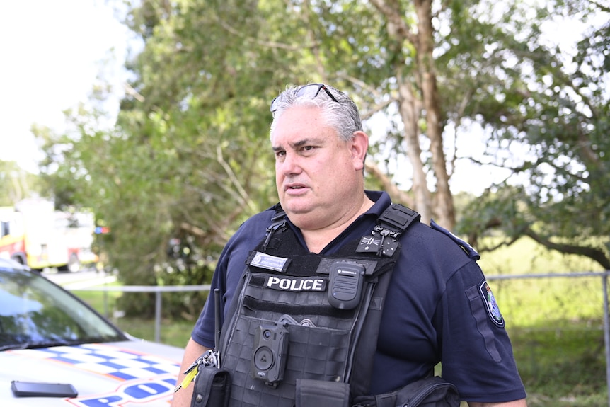 A man with grey hair in a navy Queensland police uniform stands next to a police car with trees behind him.