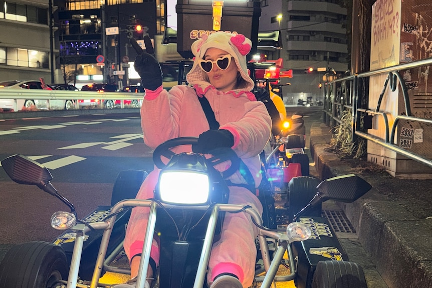 A woman in a bright pink onesie and yellow love-heart sunglasses sits in a go kart with tall Japanese city buildings behind.