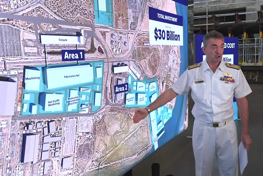 A man wearing a white navy uniform points at a map with $30 billion written on it
