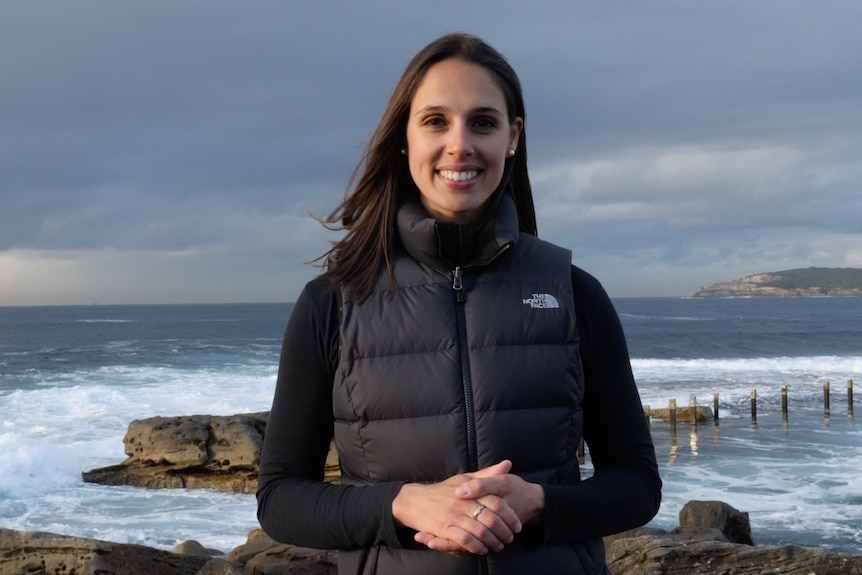 A woman stands in center frame in a black long sleeve and black vest with brown hair smiling at camera, standing at the beach.