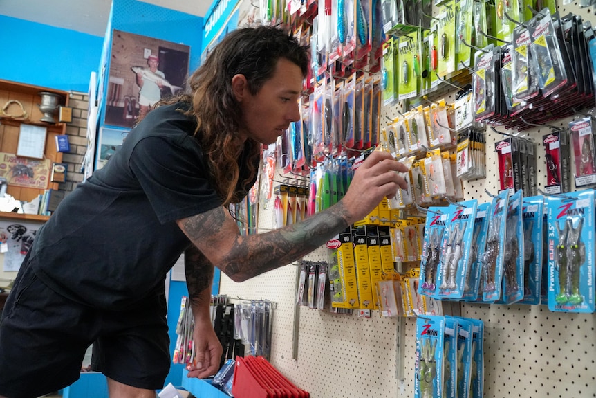 A man with long hair and tattoo stocks shelves with fishing accessories.