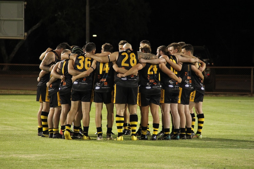 A group of men in black sporting outfits in a huddle on an oval