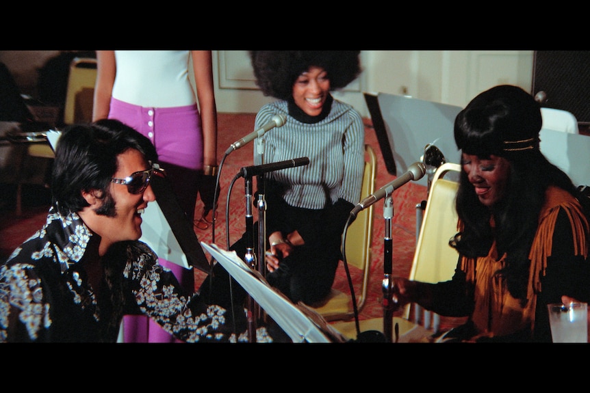 Elvis Presley sitting down laughing in a recording studio, with female African American back up singers