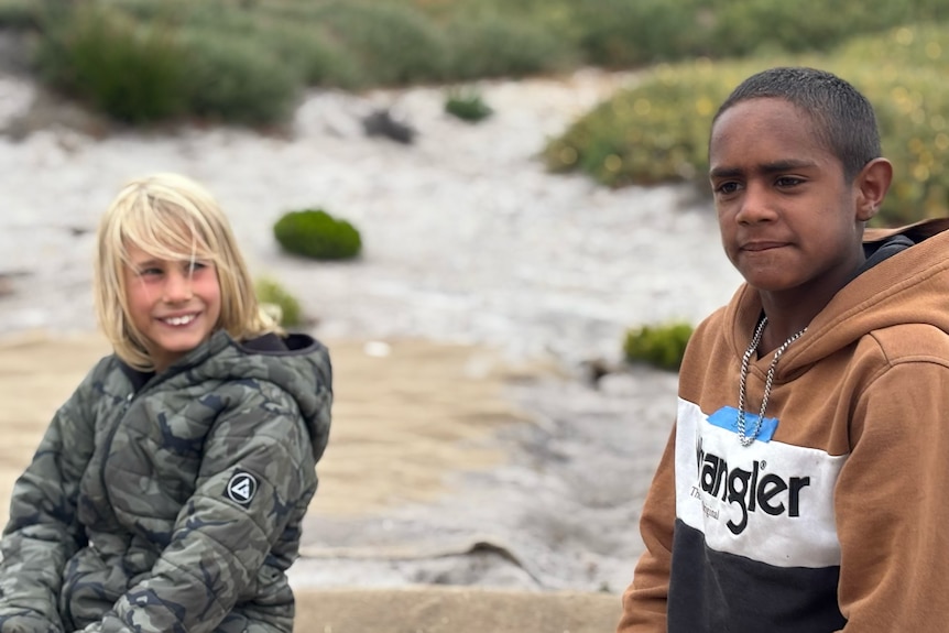 Two children sit on a sandy area near the dunes, wearing jackets and hoodies, with coastal vegetation behind them.