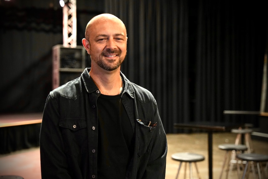 Sam wears dark clothing and has no hair. He poses for a photo inside a music theatre.