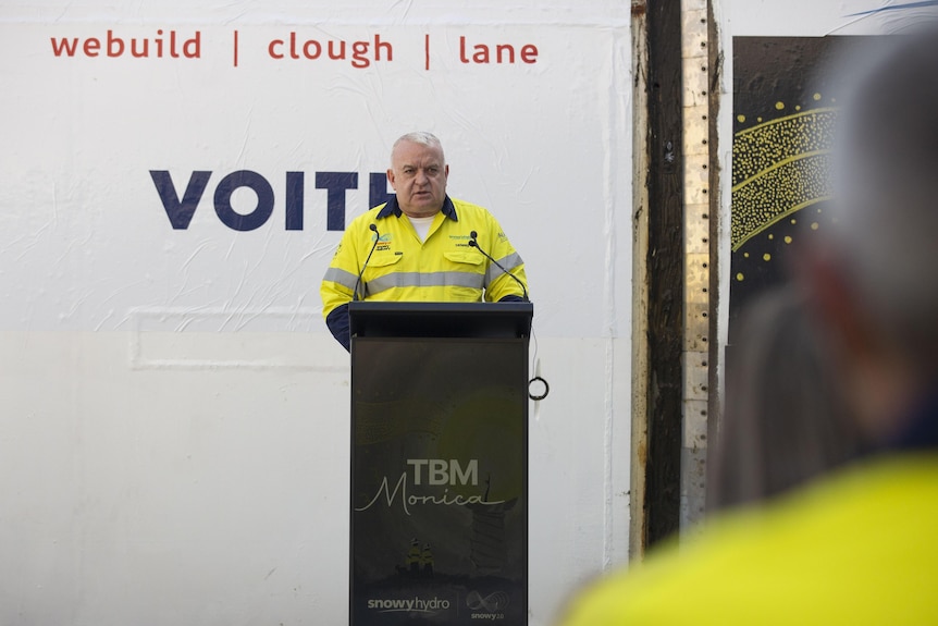 A man in a yellow construction outfit, speaking at a lecturn.