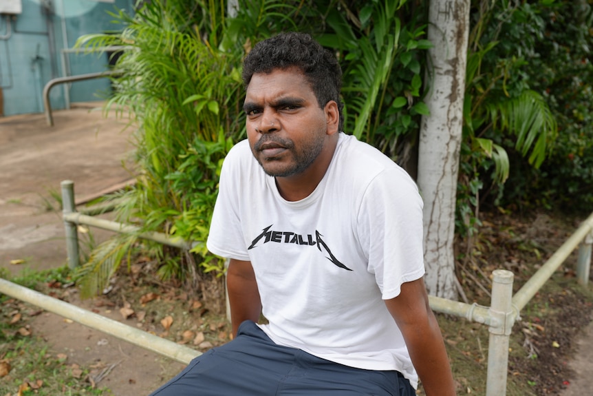 A man in a white t-shirt sits on a fence