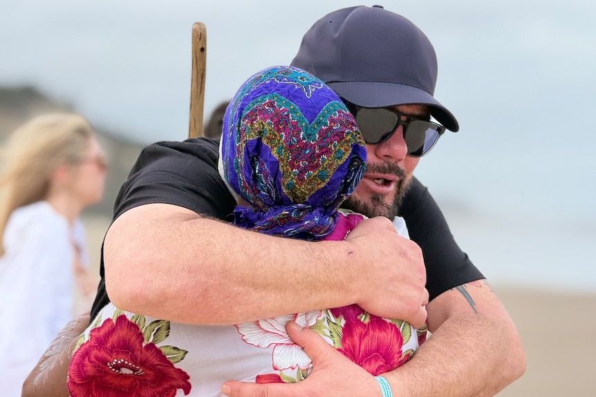 A man hugs a woman wearing a headscarf on the beach.