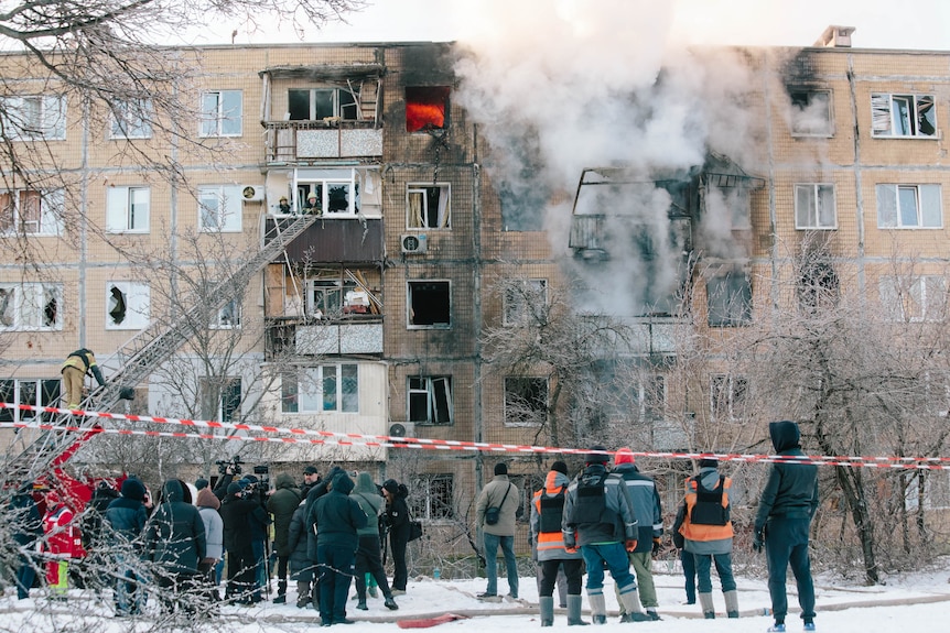 Residents in warm clothes gather in the street outside a burning building emitting smoke.