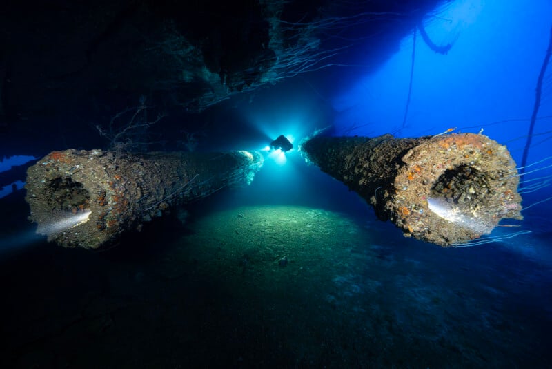 A scuba diver explores the seafloor near two large, encrusted pipes or cannons extending from a sunken shipwreck, illuminated by the diver's bright flashlight against deep blue water.