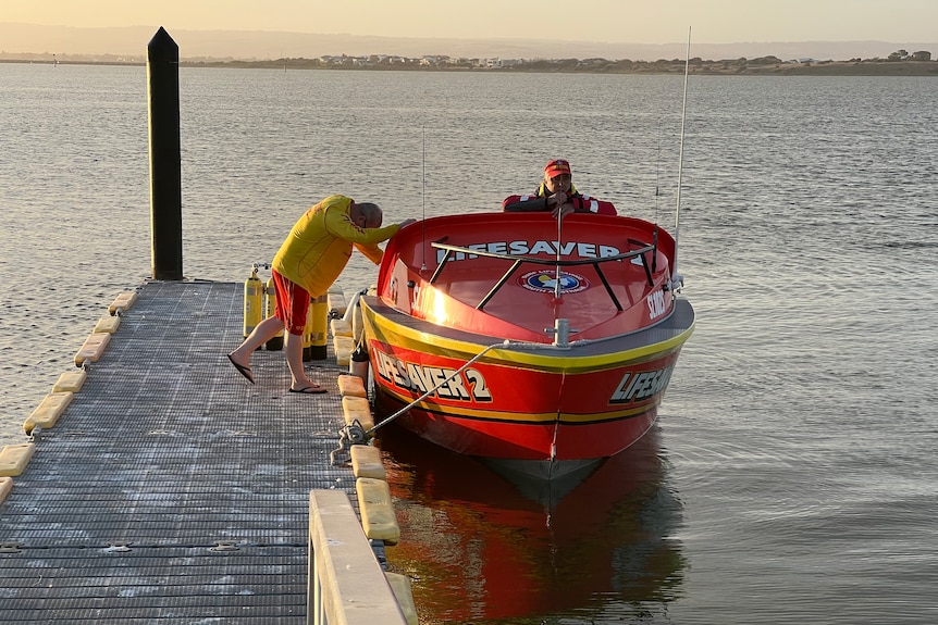A man in a yellow top and red shorts leans on a red lifesaving boat near a ramp which has a man sitting in it