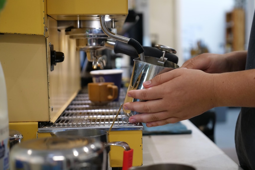 Hands holding a tin cup frothing milk on a yellow coffee machine. 