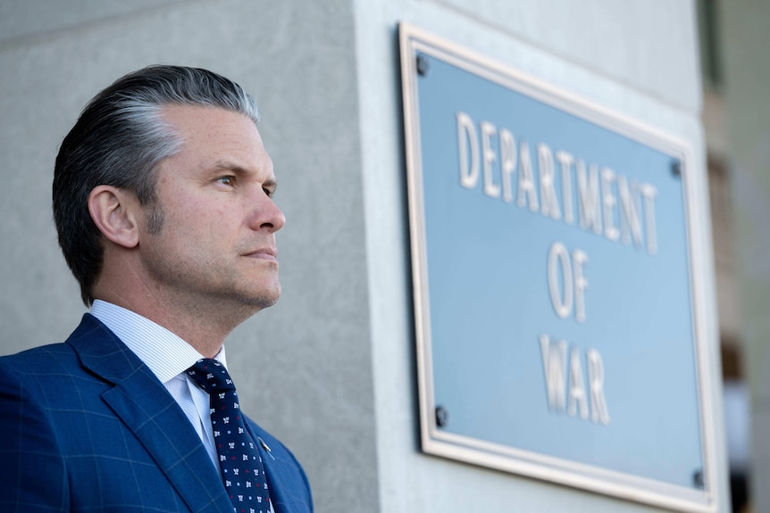 A man in a blue suit, white shirt and blue tie stands by a sign which says Department of War.
