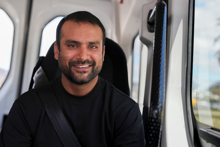 A man with dark hair and dark beard in a black t-shirt sits inside a white van with a palm tree visible through the window.
