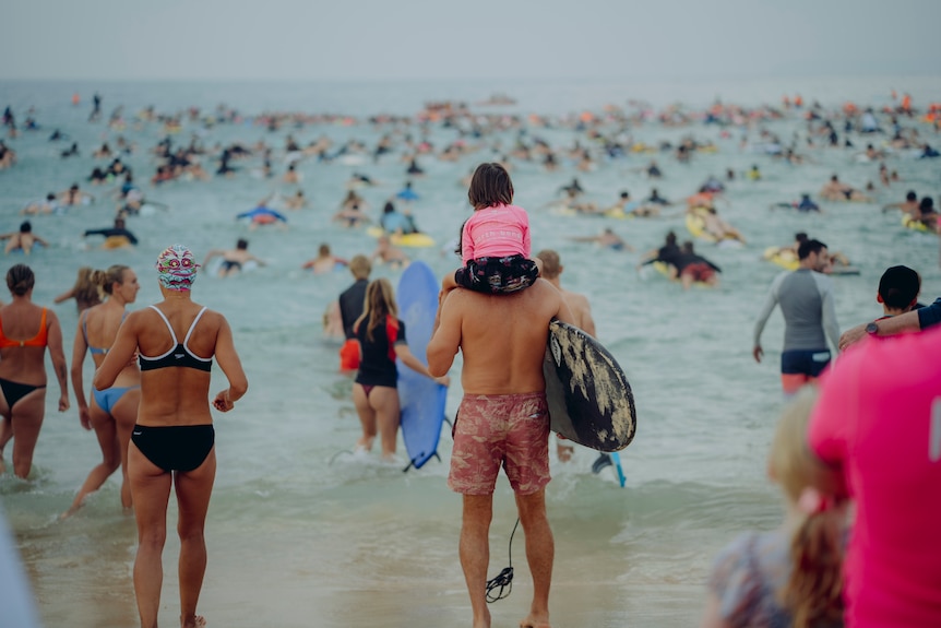 A man holding a paddle-board has his child on his shoulder as he approaches the waves where others are paddling out.