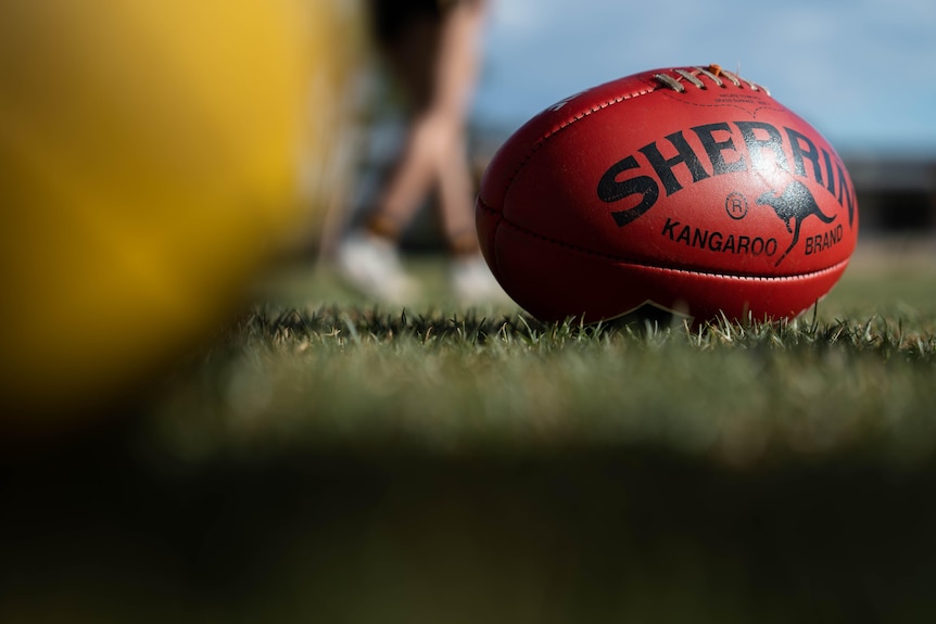 A red Sherrin Aussie Rules football on grass.