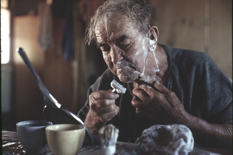 An older man looks in a mirror and shaves his face. 