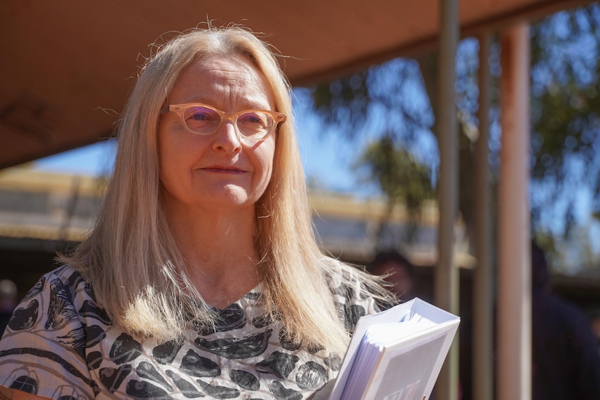 A woman with blonde long-hair, wearing pale framed glasses, holding a white folder, leaf pattern dress standing outside.