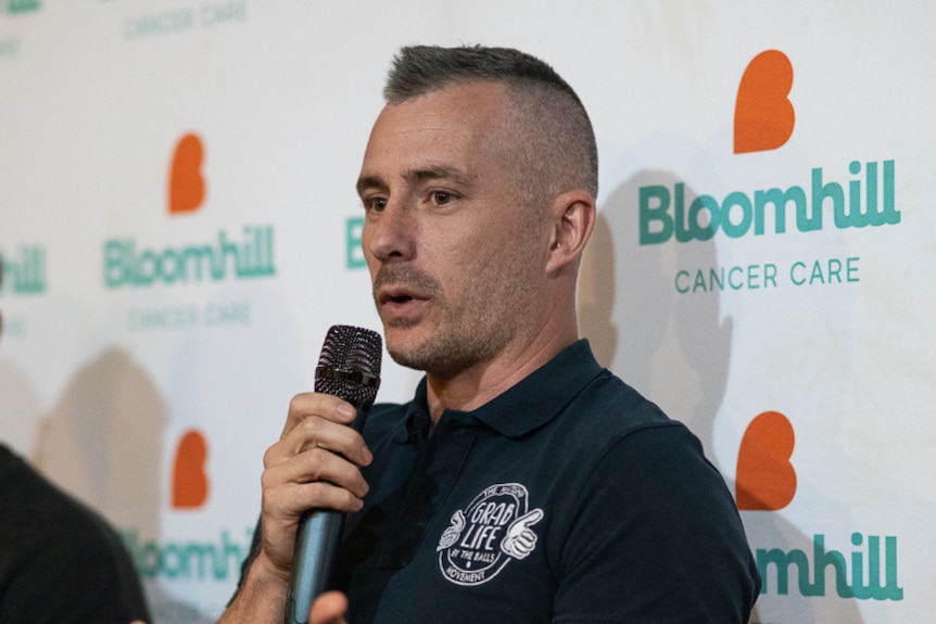 A man with a black shirt stands in front of a crowd with a microphone