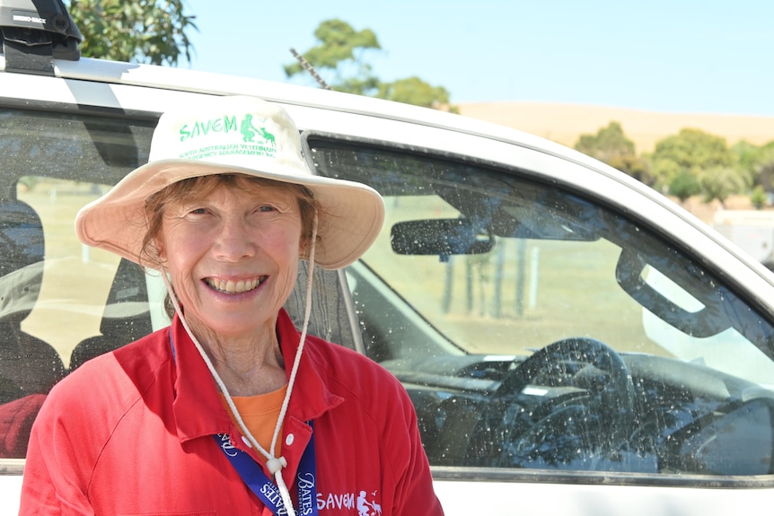 A woman in a wide brimmed hat and red overalls poses next to a vehicle
