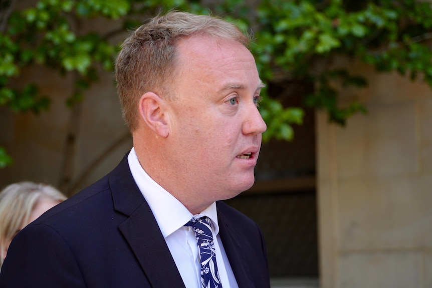 David Michael stands up at a press conference in a suit. Side on view.