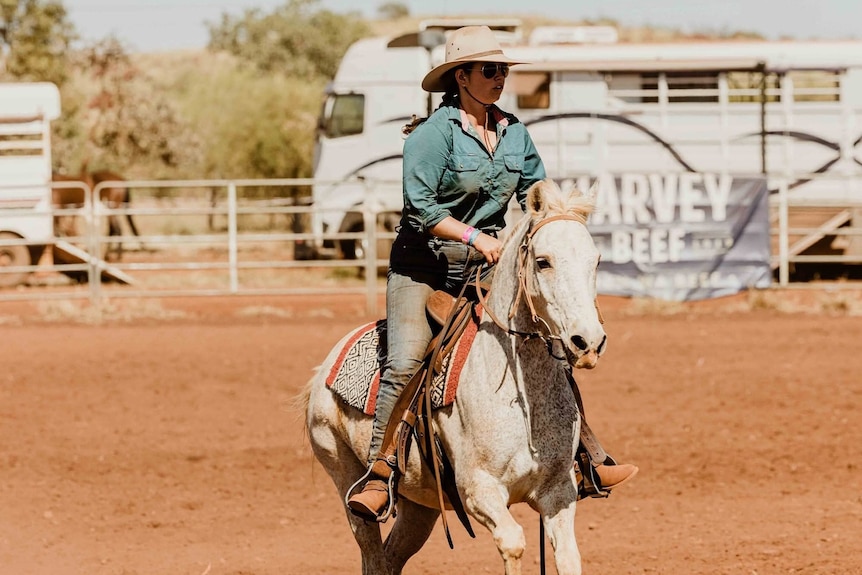 A woman in a green shirt, broadbrimmed hat, and sunglasses rides a horse in a cattle yard.
