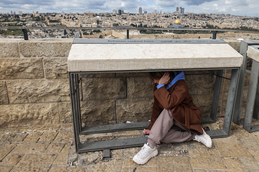 A young woman sits under an outside table covering her ears. Jerusalem can be seen in the background. 