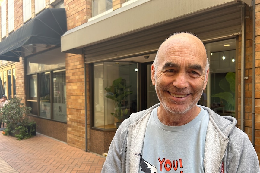 An older man in a grey hoodie smiling while standing on a street