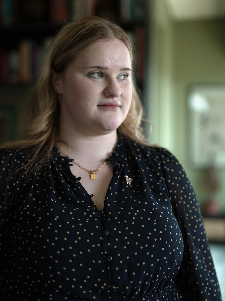 A woman stands indoors looking to the side, towards a window. She has a neutral expression on her face.