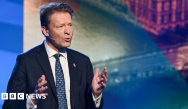 Richard Tice gestures with both hands while speaking. He's wearing a dark blue suit, and a blue tie, standing infront of a multicolored screen display.