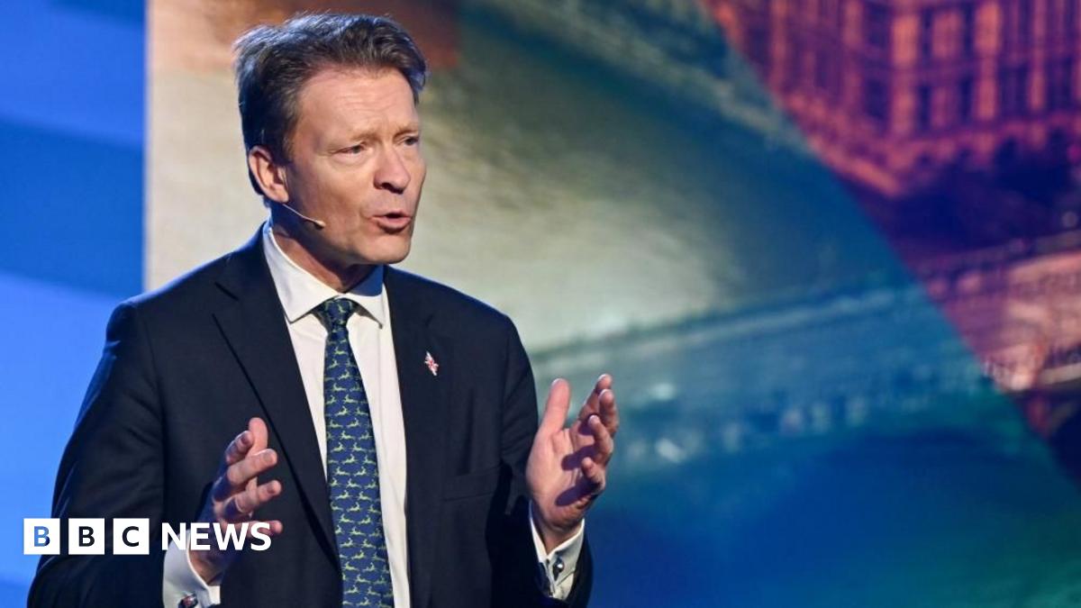 Richard Tice gestures with both hands while speaking. He's wearing a dark blue suit, and a blue tie, standing infront of a multicolored screen display.