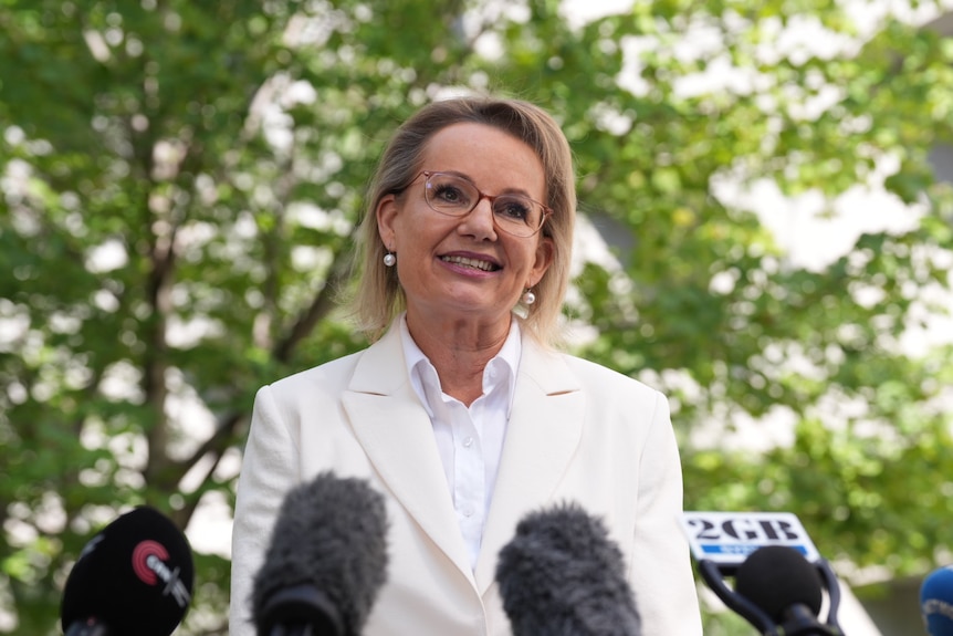 Sussan Ley, in a white suit, speaks to the media with a leafy green tree behind her