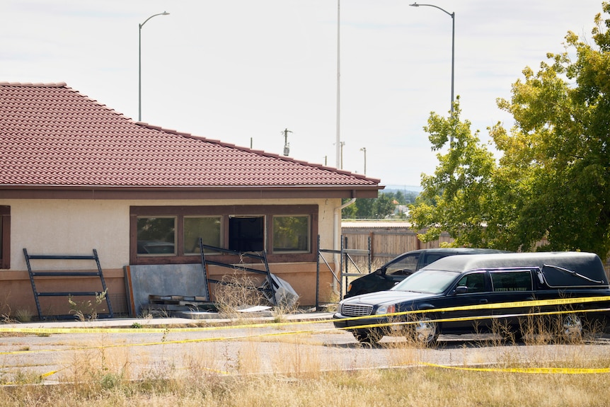 A beige funeral home with a red tile roof seen next to a black hearse and a yellow crime scene tape